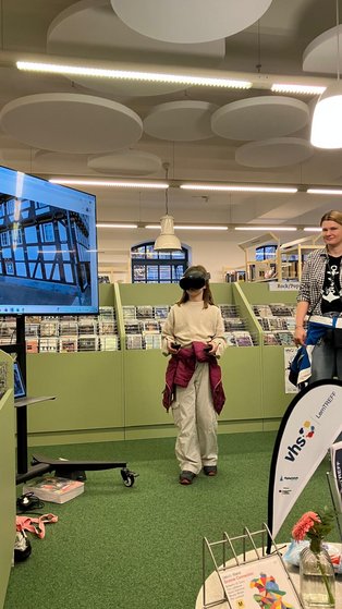 Ein Kind steht in der Mitte mit einer VR-Brille, auf einem Bildschirm kann man sehen, was das Kind durch die VR-brille sieht