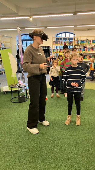 Eine junge Frau spielt mit der VR-Brille, dahinter stehen Personen, die warten