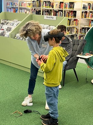 vhs-Mitarbeiterin zeigt einem Kind, das eine VR-Brille aufhat, wie das ganze funktioniert
